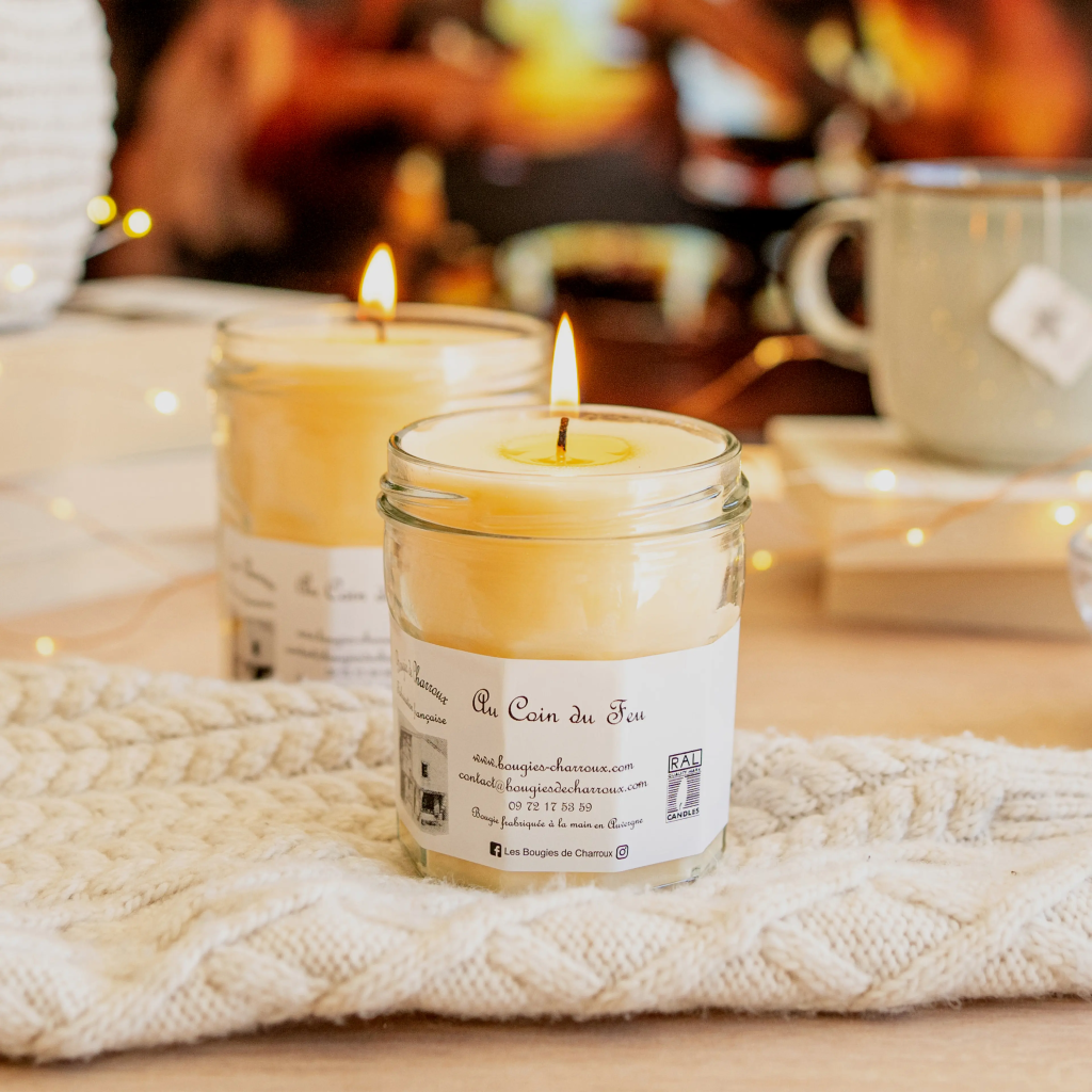 2 bougies blanches allumées dans un pot en verre. Étiquette blanche avec du texte : "au coin du feu" et leurs pseudos sur Instagram et Facebook : "les bougies de charroux". Les 2 sont dans un fond thème noël, et une ambiance chaleureuse.